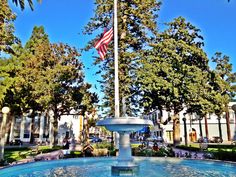 an american flag on top of a fountain in the middle of a park with people sitting and walking around