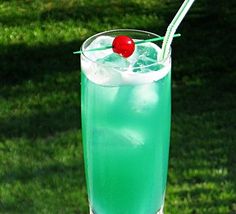 a tall glass filled with blue liquid and a cherry on the top, sitting in grass