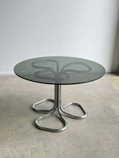 an oval glass table with metal legs and a circular design on the top, sitting in front of a white wall