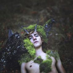 a shirtless man with horns and moss on his head is sitting in the woods