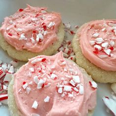 three sugar cookies with pink frosting and sprinkles on white paper next to candy canes