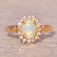 an opal and diamond ring sits on a wooden surface, surrounded by small white stones