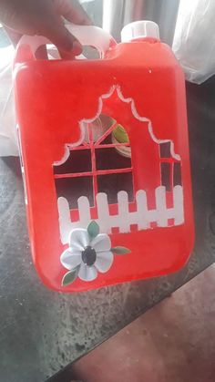 a red plastic container with white flowers and a gate painted on the side, in front of a window