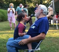 an older man sitting in a chair with a little boy on his lap and people standing around him