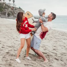 a man and woman holding a baby on the beach with a surfboard in front of them