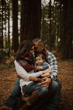 a man and woman holding a baby in their arms while sitting on the ground surrounded by trees