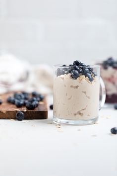 a dessert with blueberries and cream in a glass on a white table next to a cutting board