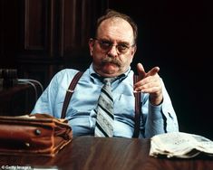 a man sitting at a table with his hand in the air and wearing suspenders