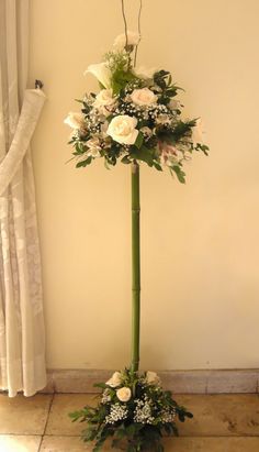 a tall vase with white flowers and greenery on the floor next to a window