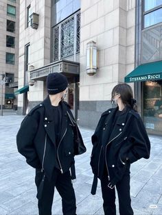 two people are standing on the sidewalk in front of a building wearing black jackets and beanies