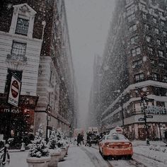 a red car driving down a snow covered street with tall buildings in the back ground