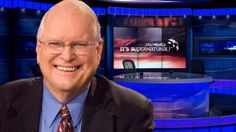 a smiling man in a suit and tie on the set of an news program with two television screens behind him