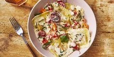 a white bowl filled with pasta and vegetables next to a glass of wine on top of a wooden table