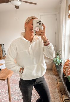 a woman taking a selfie in her living room while wearing a white sweater and jeans