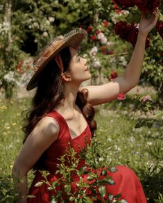 a woman in a red dress and straw hat picking roses from a bush with her hands