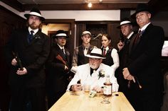 a group of men standing around a wooden table with drinks in front of them and one man wearing a hat