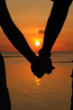 two people holding hands with the sun setting in the distance behind them, over water