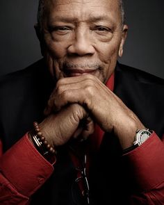 an older man wearing a red shirt and black vest with his hands on his chin