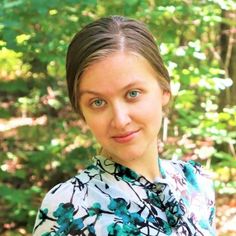 a woman standing in front of some trees and bushes wearing a shirt with blue flowers on it