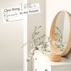 a mirror sitting on top of a white dresser next to a flower vase and sign