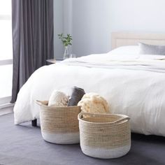 two baskets sitting on the floor in front of a bed with white sheets and pillows