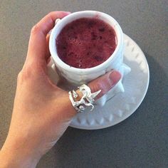 a hand holding a cup of red liquid on a white saucer with a spoon in it