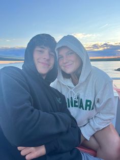 two people are sitting on a boat in the water at sunset or dawn, one is wearing a hoodie and the other is smiling