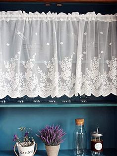 a shelf with some flowers and jars on it next to a window sill that has lace curtains over the top