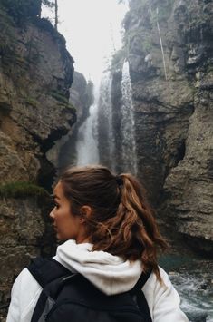 a woman standing in front of a waterfall with her back to the camera and looking at it