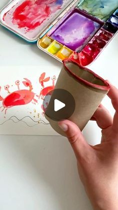 someone is holding a paper cup with paint and watercolors on the table in front of them