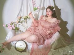 a woman in pink dress sitting on a bed with flowers and a clock next to her