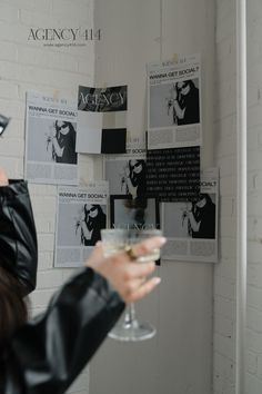 a woman holding a wine glass in front of a white brick wall with posters on it