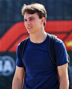 a young man with a blue shirt and black backpack smiles at the camera while standing in front of a red sign