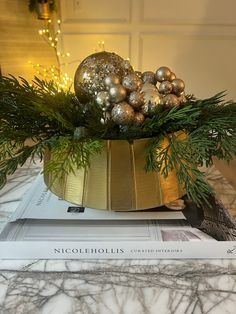 a christmas arrangement in a gold bowl on top of a stack of books with lights behind it