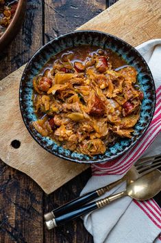 a bowl filled with meat and vegetables on top of a wooden cutting board next to silverware