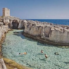 people are swimming in the clear blue water near some rocky cliffs and an old tower