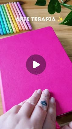 a person is writing on a notebook with colored pencils next to it and a pink cover