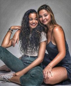 two beautiful young women sitting next to each other on top of a white floor with their arms around each other