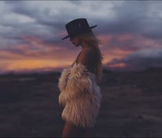 a woman wearing a hat and fur coat standing in the middle of a field at sunset