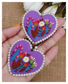 two purple heart shaped brooches with flowers on them are held in someone's hand