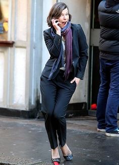 a woman walking down the street talking on her cell phone while wearing a black suit and purple scarf