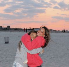 two girls hugging each other on the beach at sunset with people walking in the background