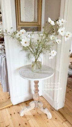 white flowers in a vase sitting on a small table next to a mirror and wooden floor