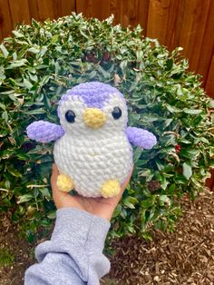 a hand is holding a small crocheted stuffed penguin in front of a bush