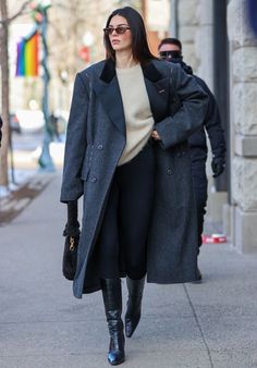 a woman walking down the street wearing black boots and a gray coat with her hands in her pockets