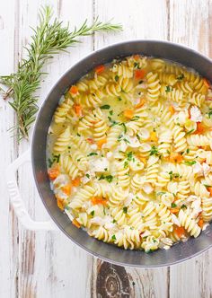 a pot filled with pasta and vegetables on top of a white wooden table next to a sprig of rosemary