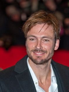 a man in a black jacket and white shirt smiles at the camera while standing next to a red carpet