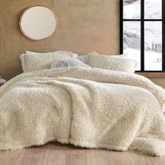 a bed covered in white fluffy blankets next to a window with a round mirror on the wall