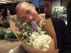 a man holding a bouquet of white and purple flowers in front of his face while looking at the camera