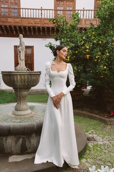 a woman standing in front of a fountain wearing a long white dress with puffy sleeves